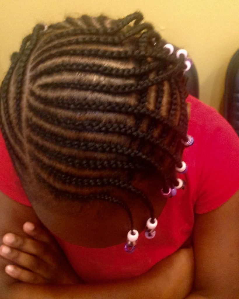 A person with beautifully braided cornrows adorned with white and purple beads on the ends, wearing a red shirt, seen from above with arms crossed.