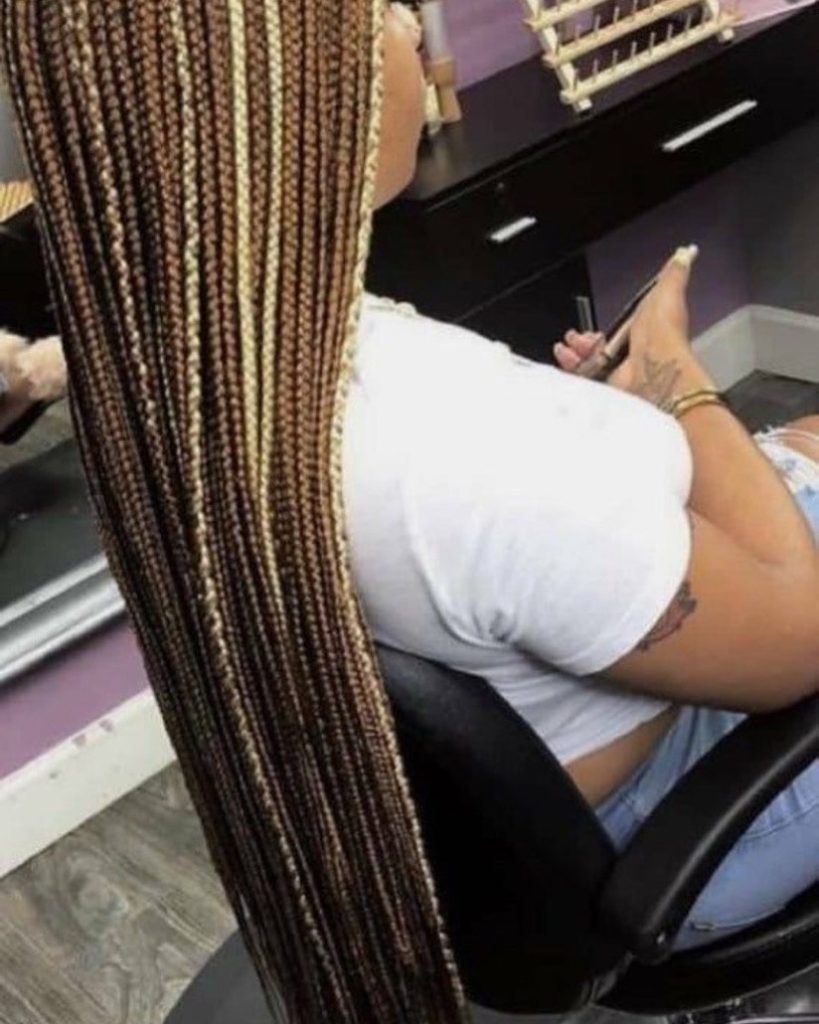 A woman with long box braids, showcasing a blend of brown and blonde, sits in a salon chair while styling her hair.