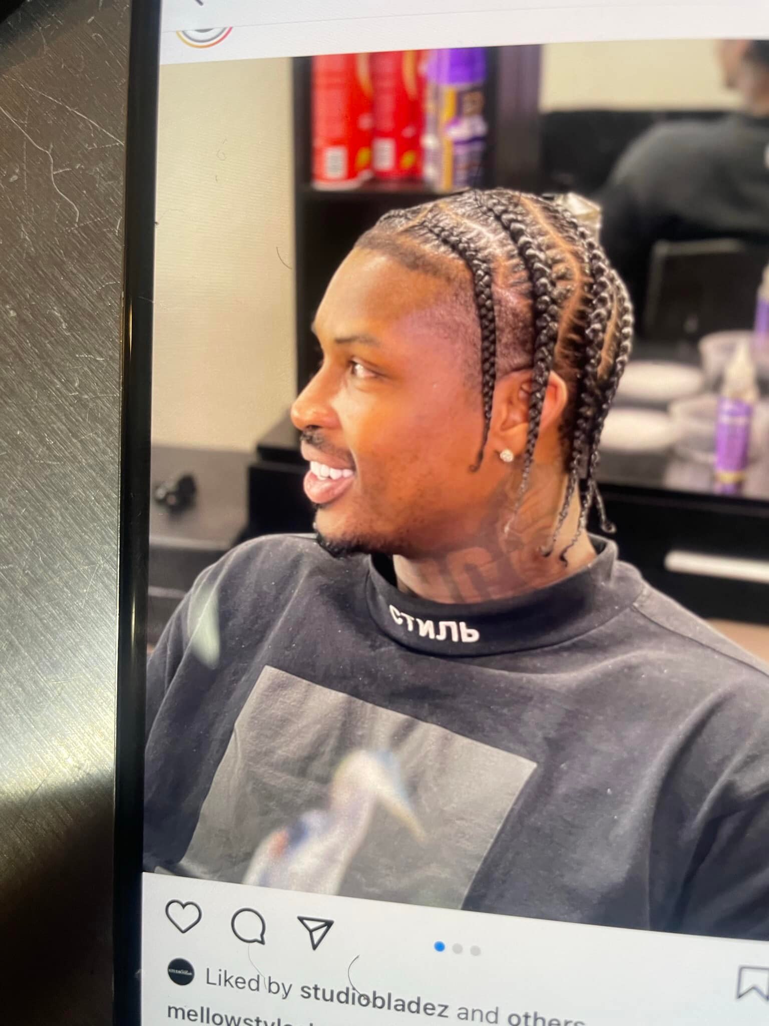 A person with braids smiles while seated in a barbershop. Shelves with hair products are visible in the background, creating a casual atmosphere.